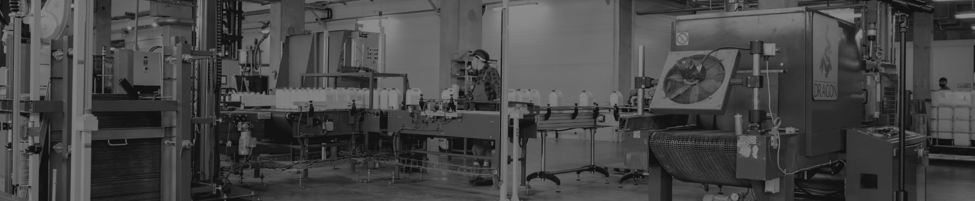 Czarno-białe zdjęcie wnętrza hali produkcyjnej firmy Dragon. Widoczne są zautomatyzowane linie produkcyjne z produktami, maszyny przemysłowe z logotypem firmy oraz pracownik w kasku ochronnym.